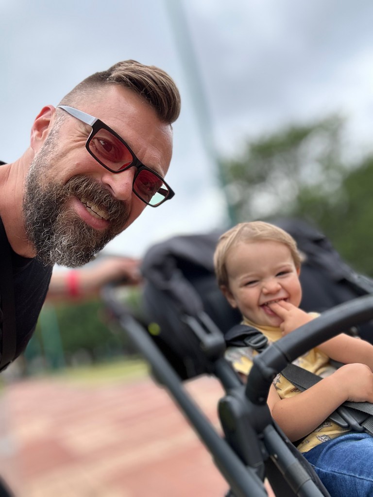 A smiling adult male with a beard and glasses poses beside a young child in a stroller outdoors, both looking happy.
